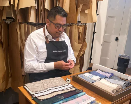 Tailor wearing Luca Falcone apron working with fabric samples in a workshop filled with hanging patterns.
