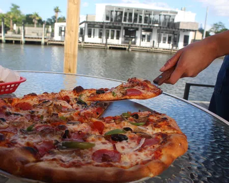 Person lifting a slice of pepperoni pizza from a glass table by the waterfront with a house in the background