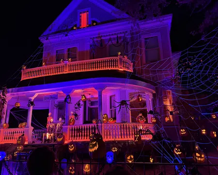 Large Victorian house decorated with glowing jack-o'-lanterns, skeletons, giant spiders, and web lights for Halloween night.