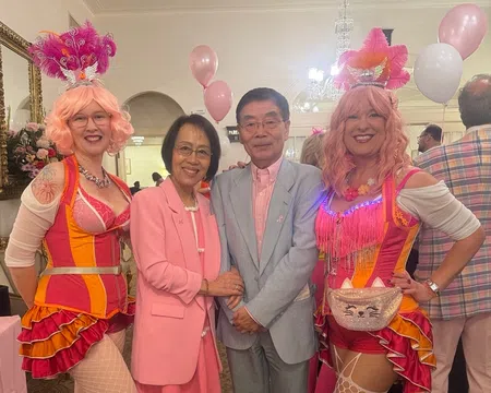 Two women in colorful circus costumes and pink wigs pose with a couple dressed in pastel pink and gray at a festive event.