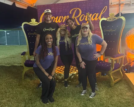 Five people wearing Crown Royal shirts pose in front of a Crown Royal backdrop between two large decorative royal chairs.