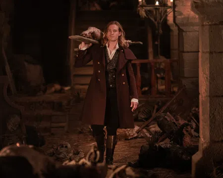 Man in historical coat carrying a wooden board with a dead animal in a dimly lit stone room with skeletons.