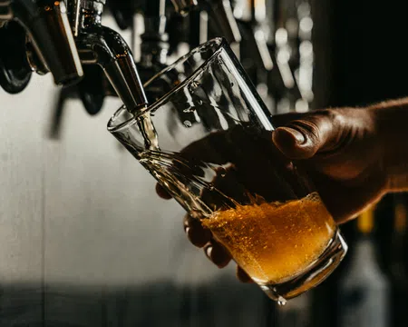Hand pouring amber beer from tap into tilted glass held by hand in dimly lit bar setting.