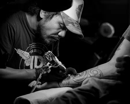 Black and white photo of a tattoo artist focused on inking a geometric tattoo on a client's forearm in dim light.
