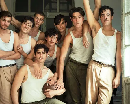 Group of eight young men wearing white tank tops and beige pants posing closely in a vintage room near open windows
