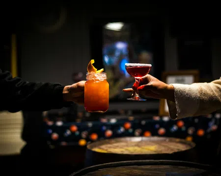 Two hands holding up colorful cocktails in dimly lit bar with wooden barrels in the foreground.