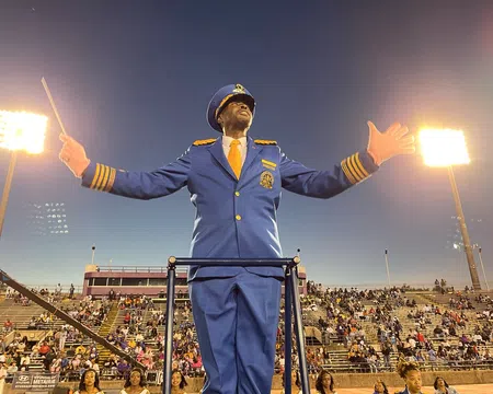 Band director in blue uniform conducting with baton at a stadium filled with spectators during evening.
