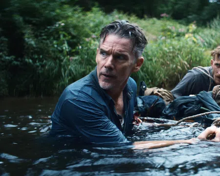 Two men cautiously wading through a river surrounded by dense greenery, holding onto a rope for support.