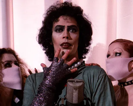 Actor in iconic glam rock makeup and costume with two masked fans on either side against a pale curtain.