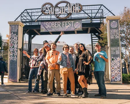 Group of people posing and smiling under Sober Fest banner at outdoor event entrance on sunny day