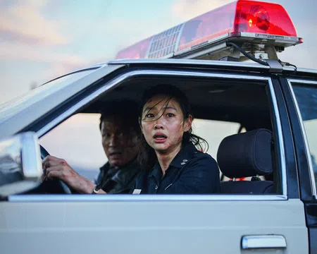 Female police officer and male colleague in a police car with red siren light, looking concerned through open window.