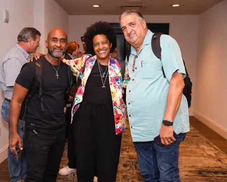 Three diverse adults smiling and posing together indoors at a social event in a hallway.