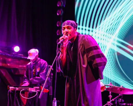 Hip-hop artist performing on stage with microphone and DJ in the background under vibrant purple and blue lights.