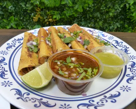 Plate of golden taquitos garnished with cilantro and onions, served with lime wedges and two dipping sauces.