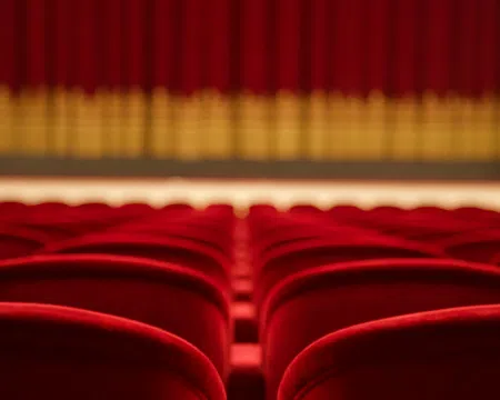 Empty red theater seats facing a closed stage curtain with golden accents under warm lighting.