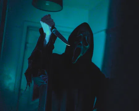 Person wearing a ghost face mask and dark robe holding a raised knife in a dimly lit room.