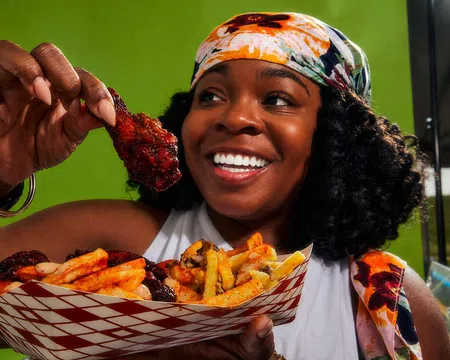 Smiling woman holding a saucy chicken wing and a tray of seasoned fries against a green background.