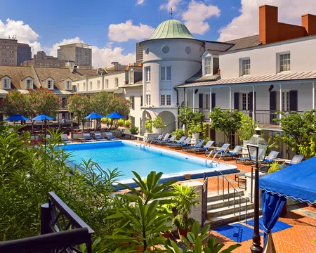 Outdoor pool area surrounded by lounge chairs, greenery, and historic white buildings under a blue sky.