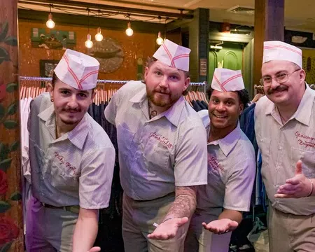 Four men wearing retro diner uniforms and hats posing with outstretched hands in a cozy restaurant setting.