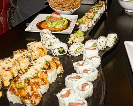 Variety of sushi rolls arranged on a black table with sauces, avocado, and garnishes in a restaurant setting.