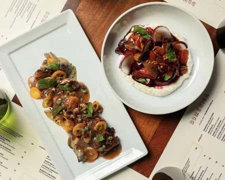 Gourmet dishes with sauces and herbs served on white plates surrounded by menus and wine glasses on wooden table.