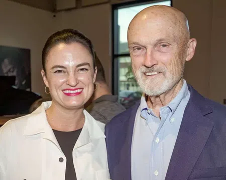 Smiling woman and older man standing indoors near a window in a casual social setting.