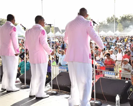Five male singers in pink jackets performing on outdoor stage before large audience at daytime event.