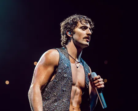 Muscular male singer with curly hair and sleeveless studded vest performing on stage with microphone