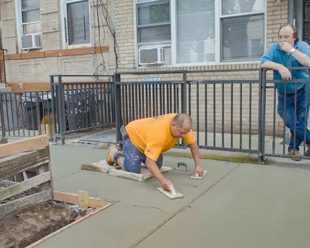 Construction worker smoothing fresh concrete on a sidewalk while another man watches at a residential building.