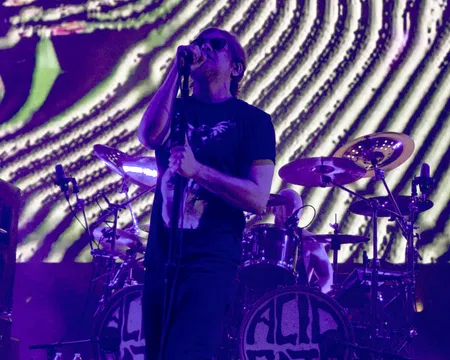 Male rock vocalist singing on stage with microphone and drummer performing behind during purple light concert.