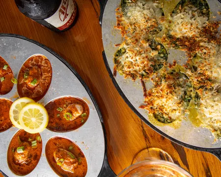Baked oysters with cheese and spinach alongside oysters in spicy red sauce garnished with lemon slices on wooden table.