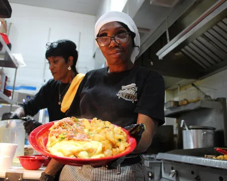 Smiling chef in kitchen holding a red plate with breakfast food including eggs, potatoes, and bacon.