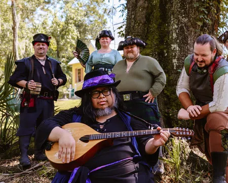 Group of six people in medieval and pirate costumes posing outdoors with a lute player in front under a large tree.