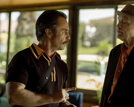Two men in conversation, one wearing a hat and suit, the other in a black shirt with orange collar details indoors near windows.