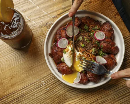Bowl of fried plantains with poached egg, shredded meat, and radish slices on a wooden table with iced tea.