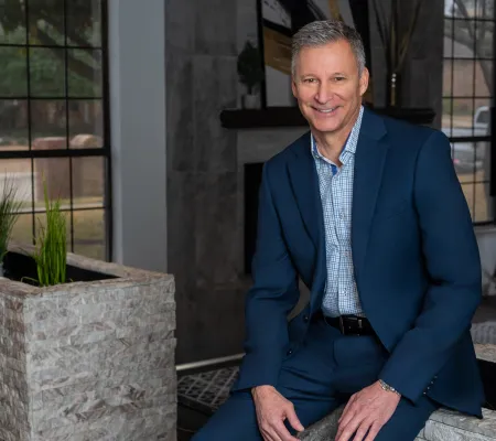Smiling middle-aged man in blue suit sitting indoors near stone planters and large windows, natural light.
