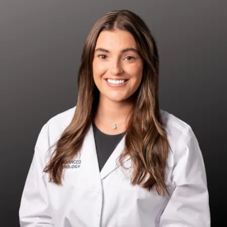 Smiling female scientist with long brown hair wearing a white lab coat against a dark gray background