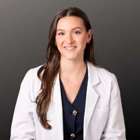 Young female doctor with long brown hair wearing a white lab coat and navy scrubs against dark background