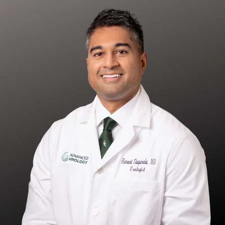 Smiling male doctor in white coat and green tie posing against dark gray background in professional headshot.