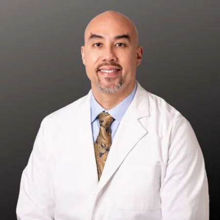 Smiling male doctor wearing a white coat, blue shirt, and patterned tie against a dark gray background.