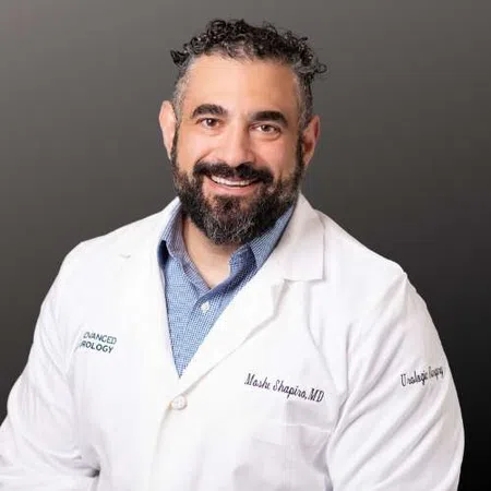 Smiling male doctor with beard wearing white lab coat and blue shirt against a neutral gray background