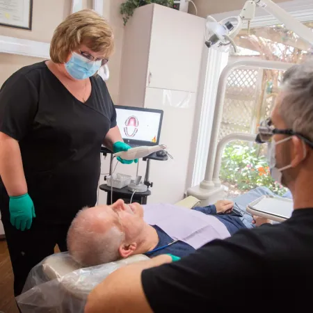 Dentist and assistant examining senior male patient in dental clinic with equipment and natural light.
