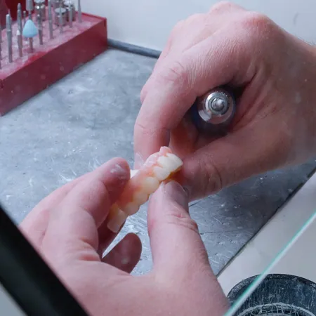 Hands polishing a dental prosthetic with a rotary tool on a workbench with dental drill bits.