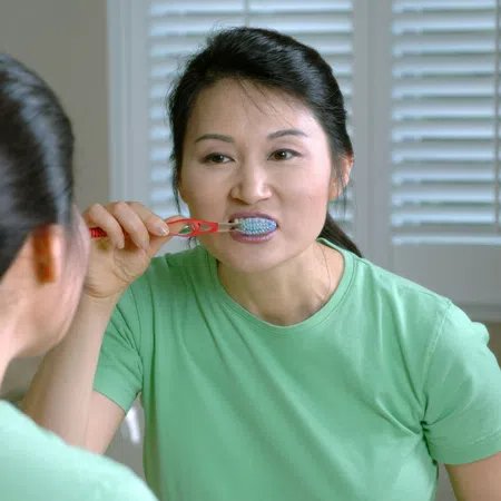 Woman in green shirt brushing teeth while looking in mirror with natural light from window blinds.