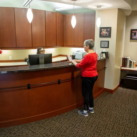 Senior woman checking in at a warmly lit medical reception with wooden desk and floral arrangement.