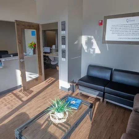 Bright dental office waiting room with black couch, glass coffee table, and reception desk with open door.