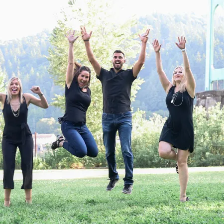 A group of six people joyfully jumping in the air outdoors, with greenery and a bridge in the background.