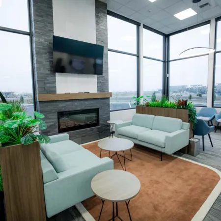 Modern office lounge with light blue sofas, round wooden tables, fireplace, large windows, and green plants.