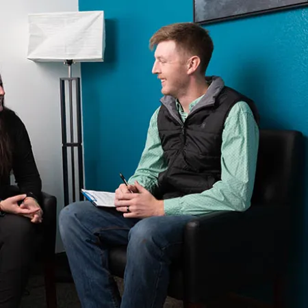 Man and woman smiling and talking while seated in black chairs in a cozy room with blue and white walls.