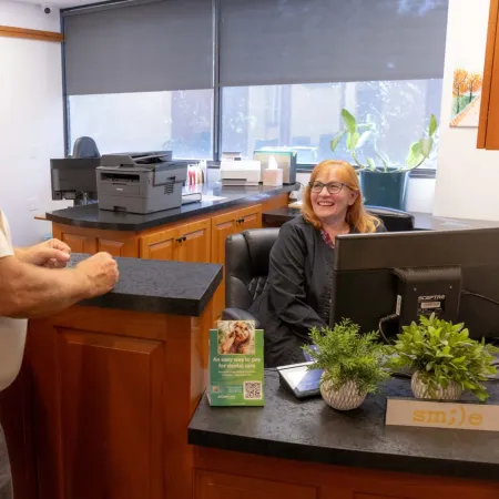 Man talking to smiling receptionist in dental office reception area with plants and informational brochure.